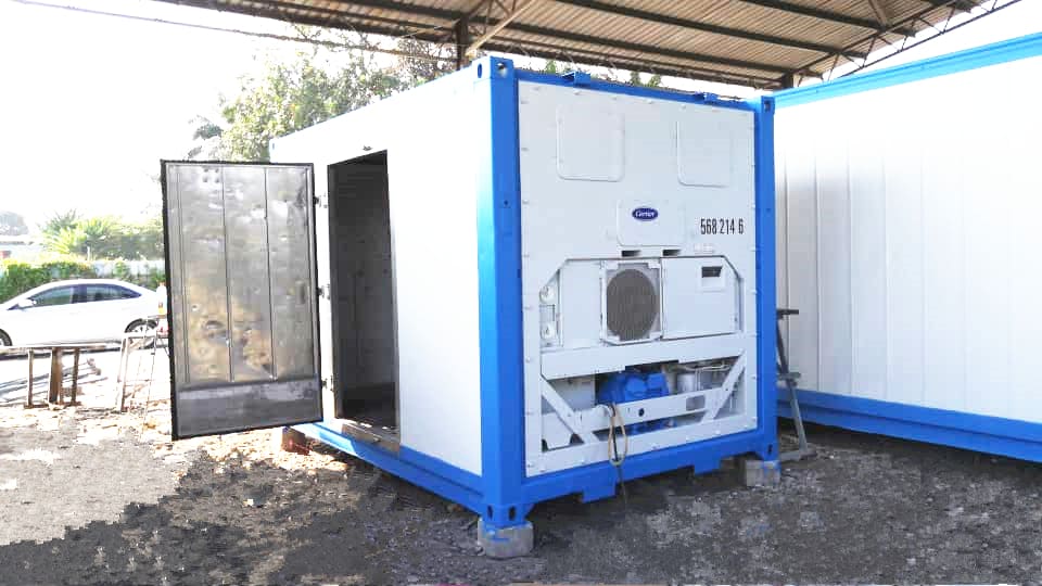 A blue industrial generator inside a container with an open door, placed next to a white building.