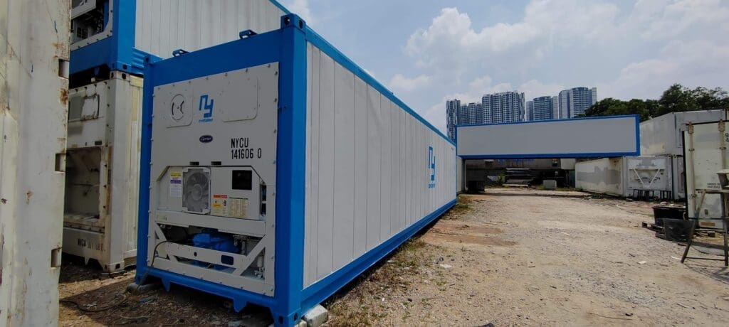 Blue reefer container converted into a mobile office or workspace, situated in an industrial outdoor area with other containers and buildings in the background.